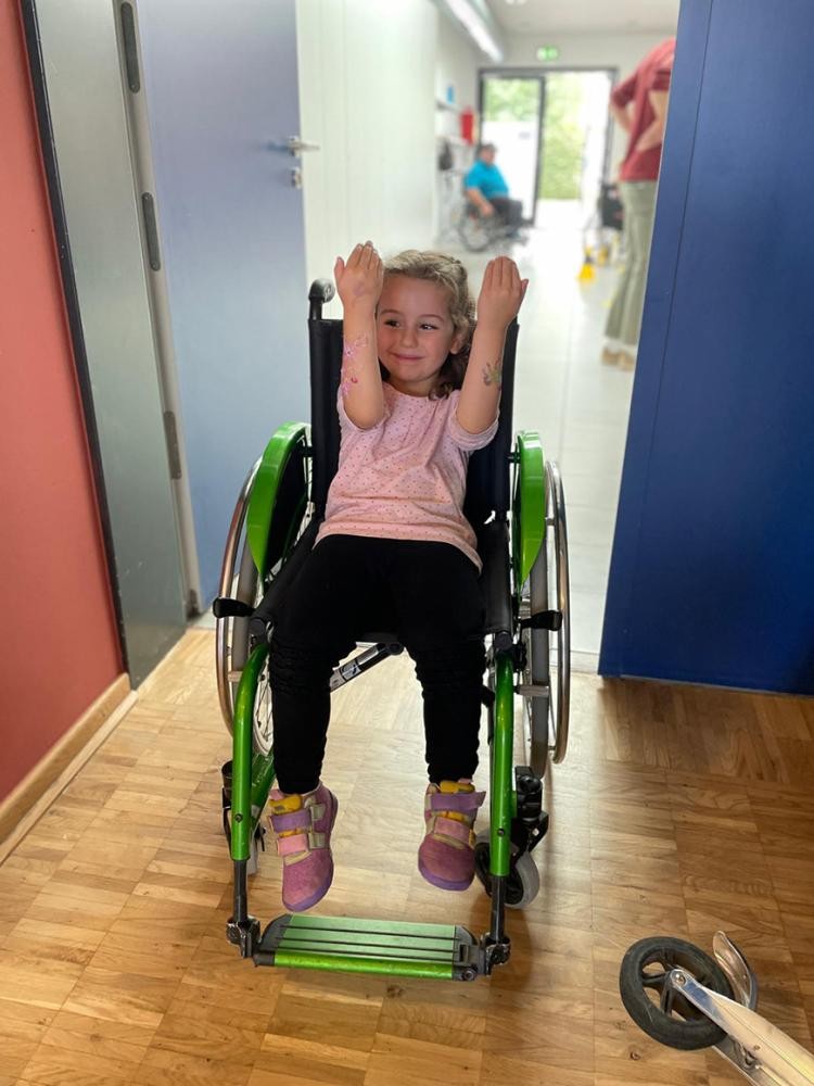 Stadtverwaltung Ein junges Mädchen sitzt in einem Gang in einem Rollstuhl und zeigt fröhlich ihre bunt gestalteten Arme.