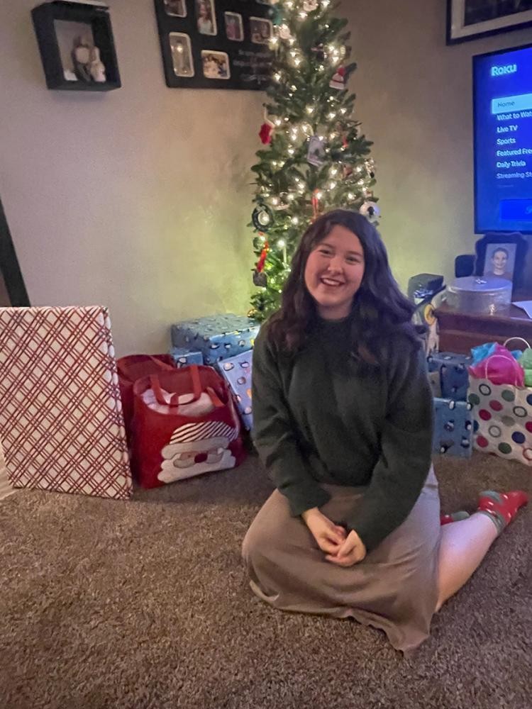 privat Eine junge Frau sitzt lächelnd vor einem Weihnachtsbaum unter dem Geschenke liegen