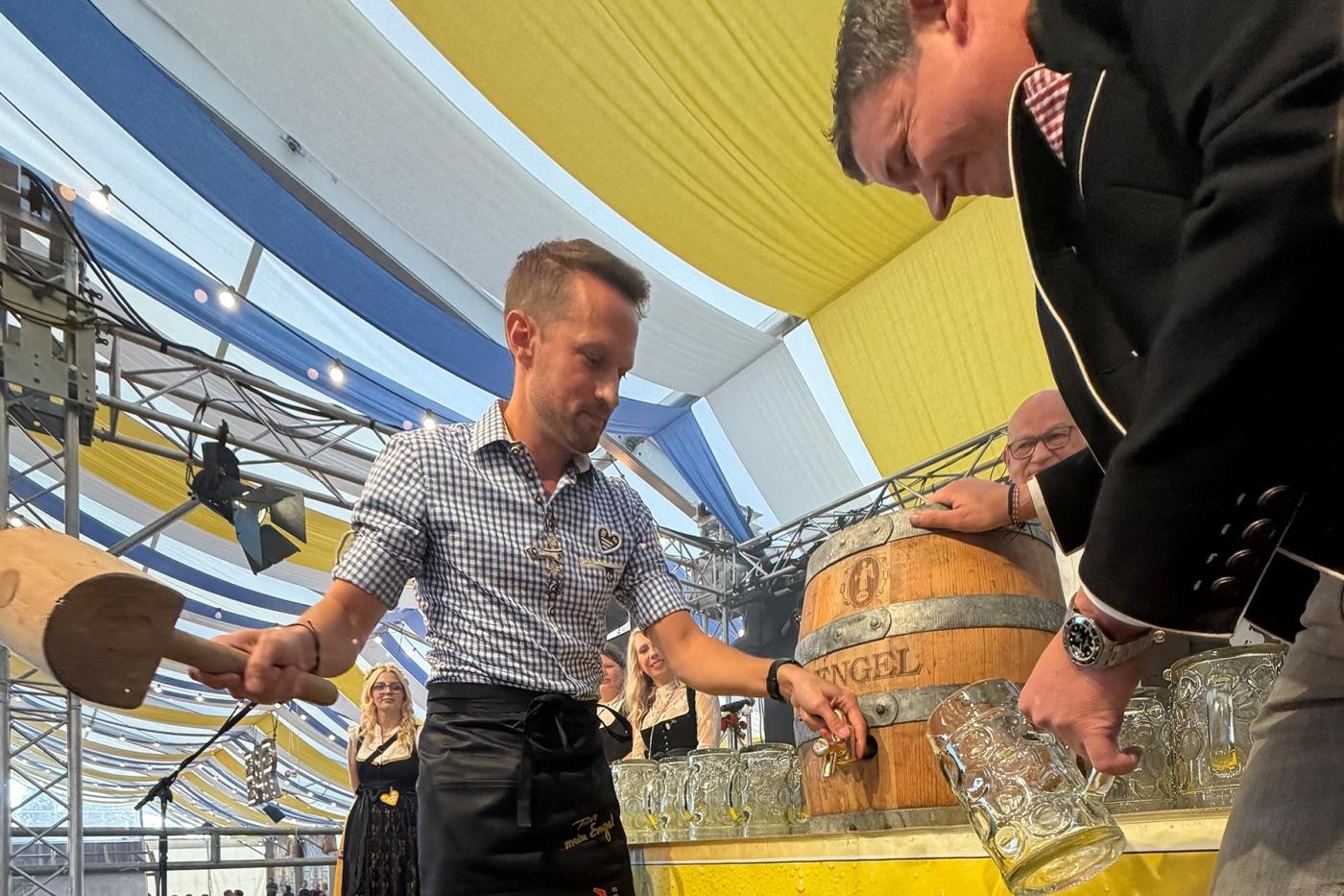 Ein Mann mit großem Holzhammer schlägt auf einen Bierhahn in einem Fass, ein anderer hält einen Krug bereit