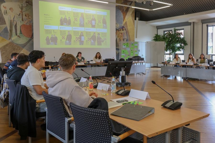 Einige junge Leute sitzen an Tischen, im Hintergrund ist eine Präsentation zu sehen.