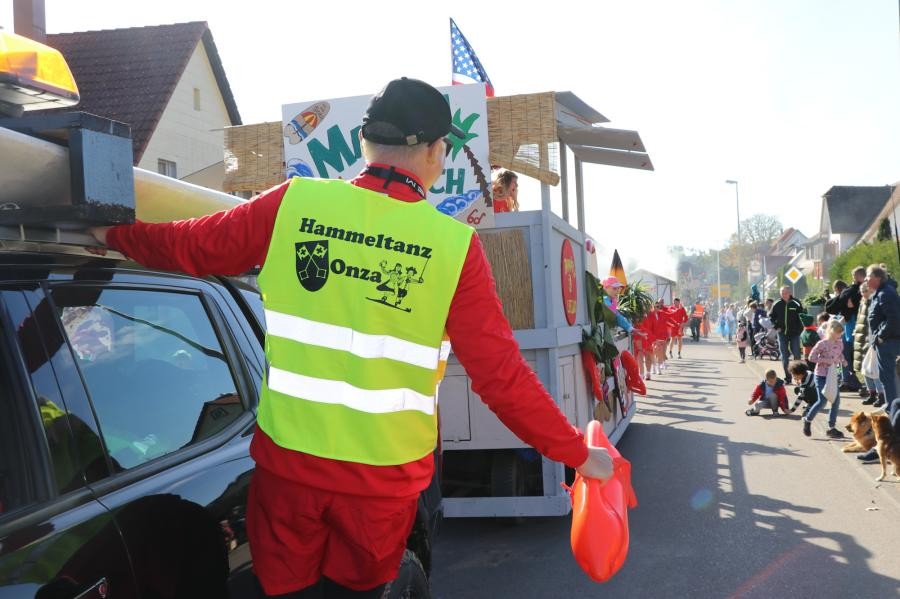 Ein Mann mit gelber Warnweste hängt an einem Auto und hat eine Rettungsboje in der Hand, dahinter ist ein Festzug zu sehen.
