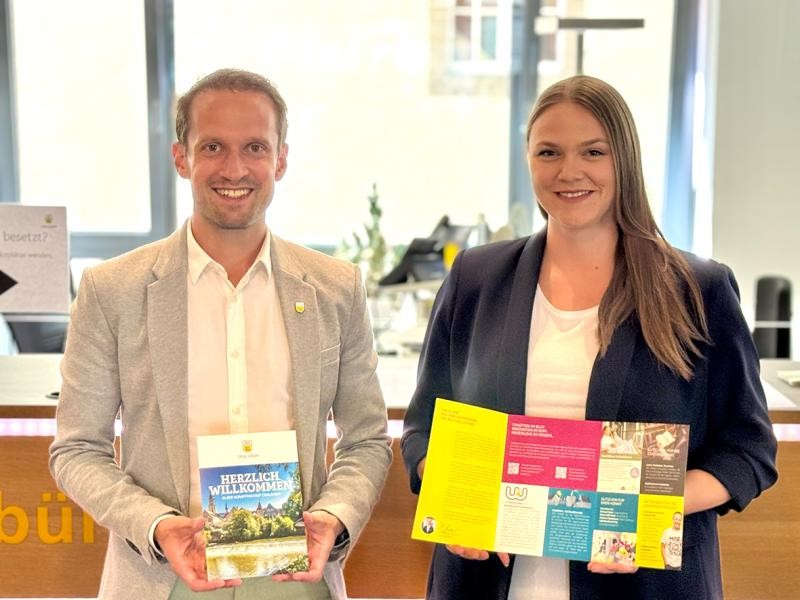 Ein mann udn eine Frau lächeln in die kamera udn halten jeweils ein Faltblatt in der Hand