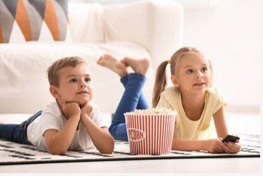 Zwei Kinder liegen vor einem Sofa auf einem Teppich und zwischen ihnen steht ein Popcorneimer
