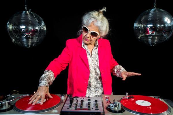 Eine ältere Dame in einem knallpinken Blazer und einer großen Sonnenbrille steht an einem DJ-Pult und legt Platten auf.