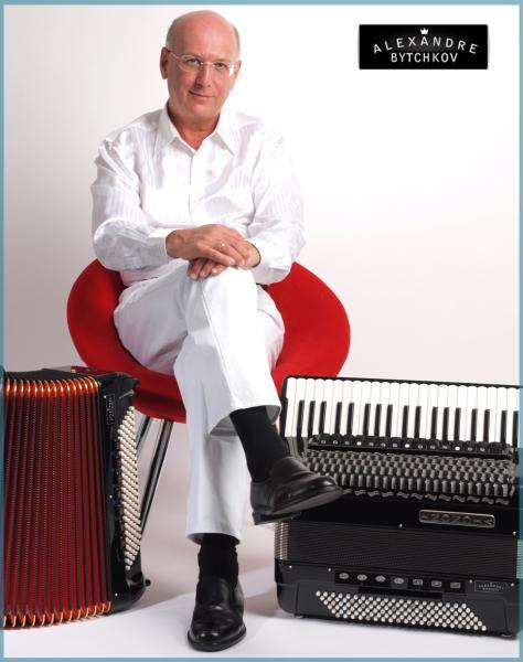 Mann in weiß sitzt auf einem roten Stuhl daneben jeweils eine Ziehharmonika