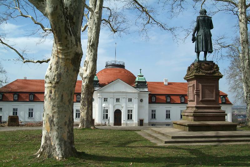 Älteres großes Gebäude mit rotem Dach, einer Statue im Vordergrund sowie ein paar Bäumen