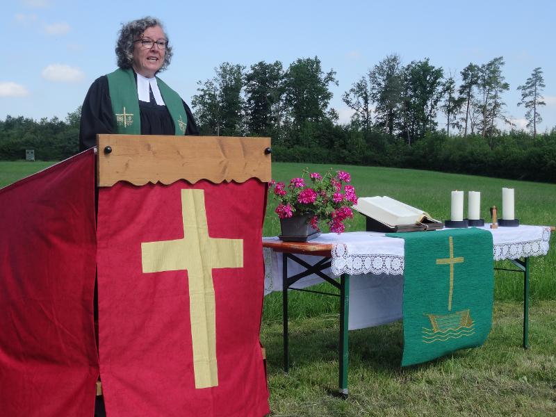 Pfarrerin hält eine Predigt auf einem Feld es ist ein Altar ähnlicher Tisch neben dran aufgebaut