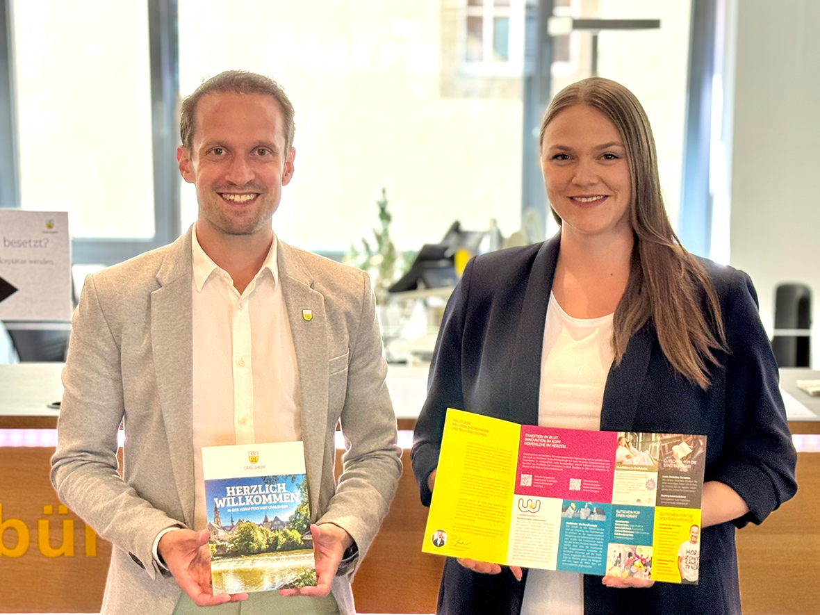 Ein mann udn eine Frau lächeln in die kamera udn halten jeweils ein Faltblatt in der Hand