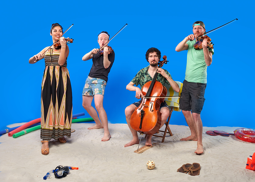 vier Musiker stehen auf einem künstlichen Strand vor einem blauen Hintergrund