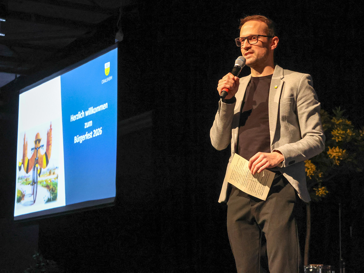 Ein Mann mit Mikrofon steht auf einer Bühne vor einer Leinwand