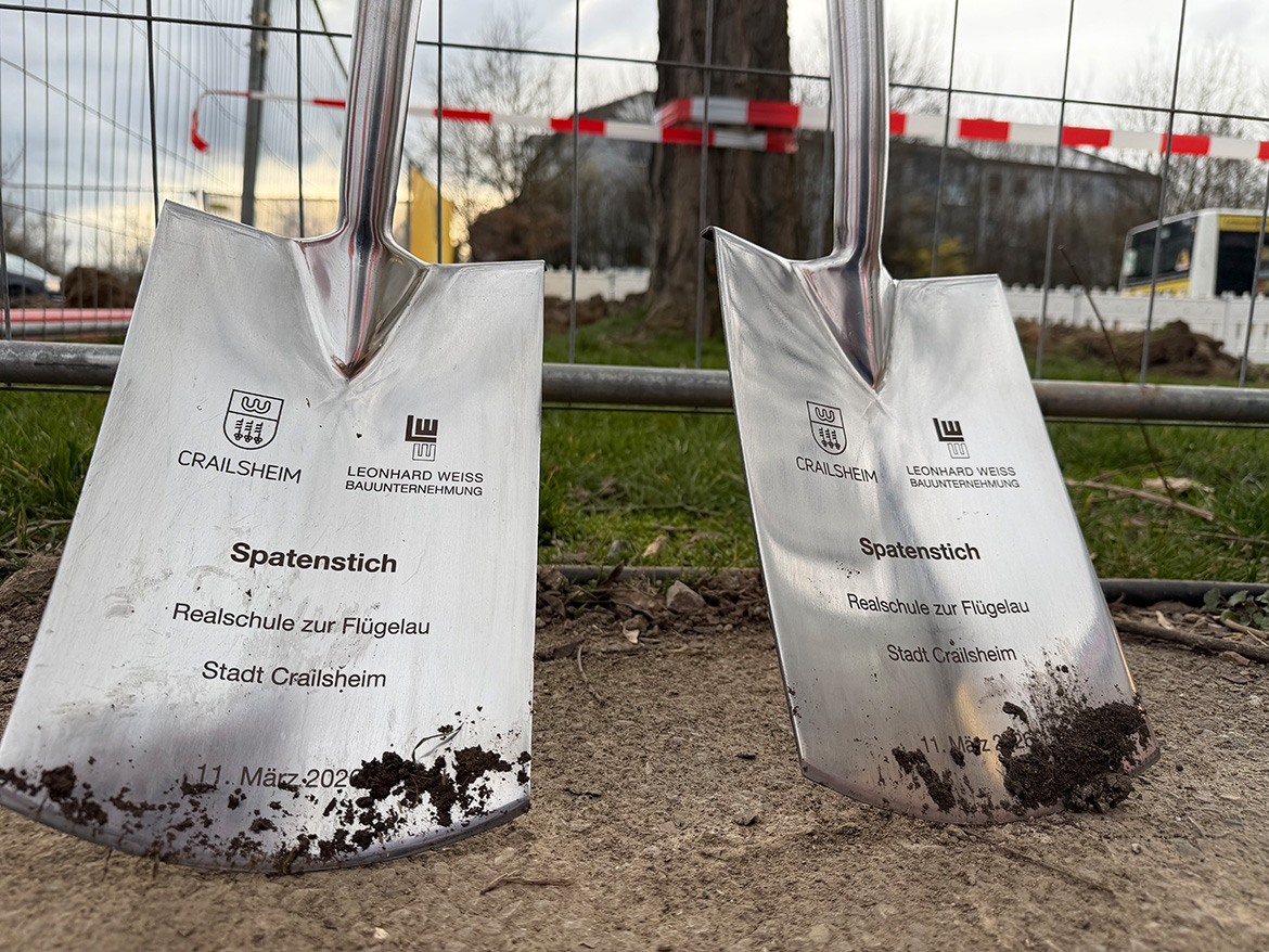 Zwei silberfarbene Spaten, auf denen ein Stadt- und ein Firmenlogo sowie ein Datum gedruckt sind