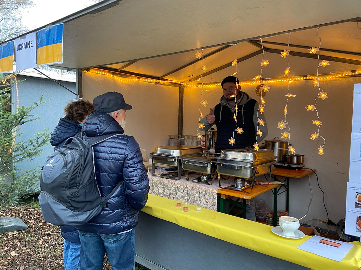 Gäste stehen vor einem Stand, dessen Betreiber in die Kamera winkt