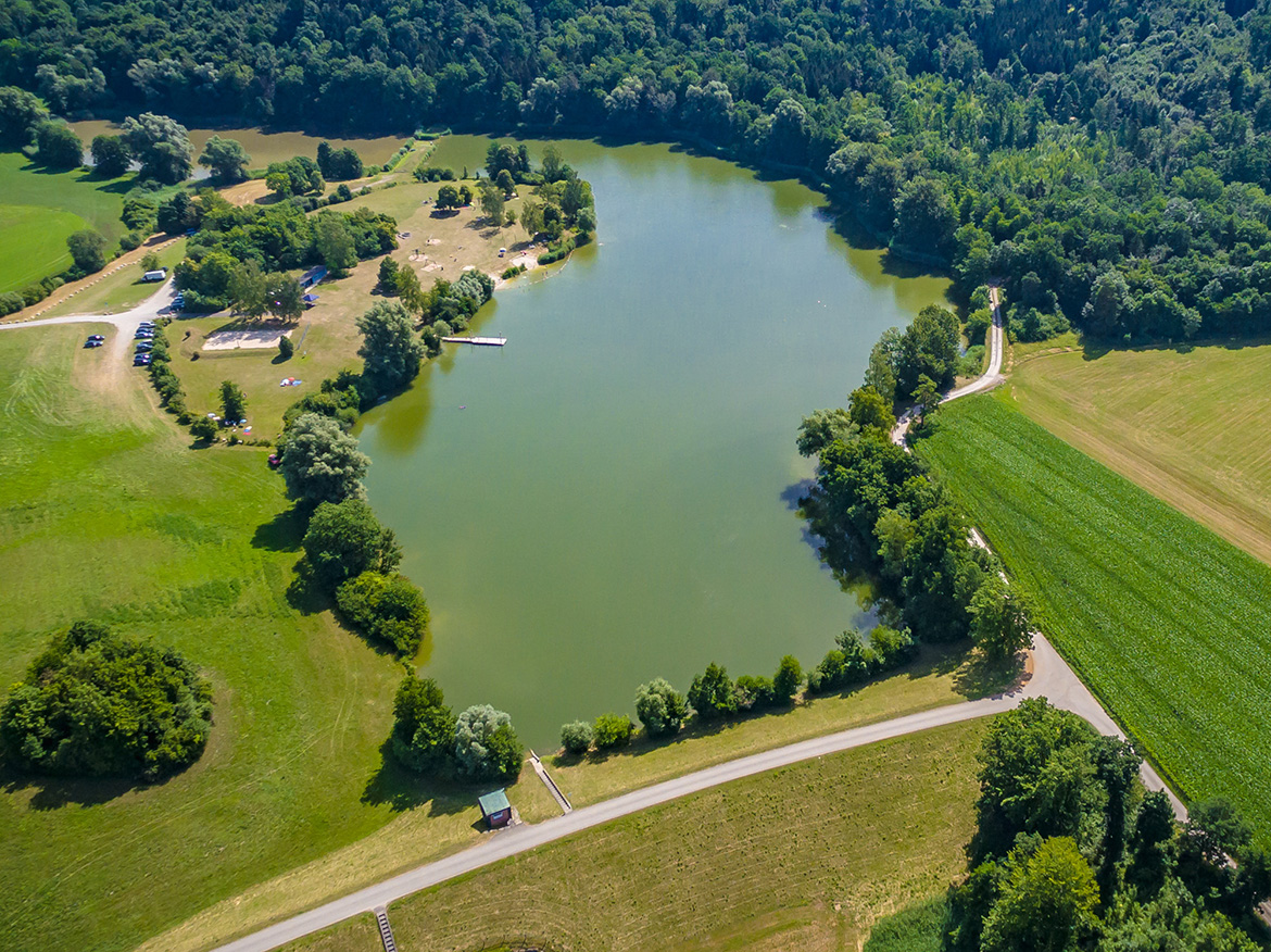 ein See aus der Vogelperspektive umgeben von Wald und Wiesen