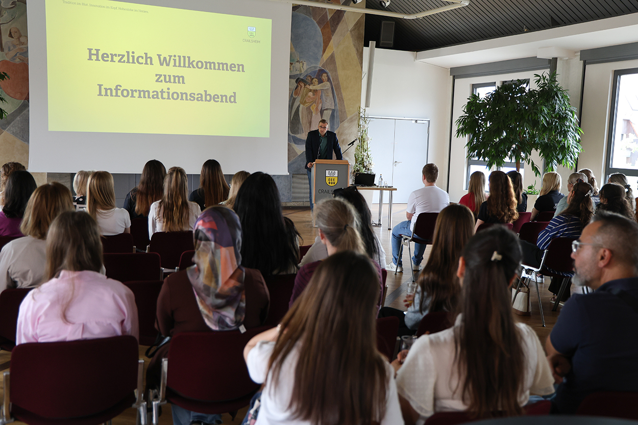 Ein Mann steht vor einer Leinwand an einem Rednerpult vor vielen sitzenden Menschen