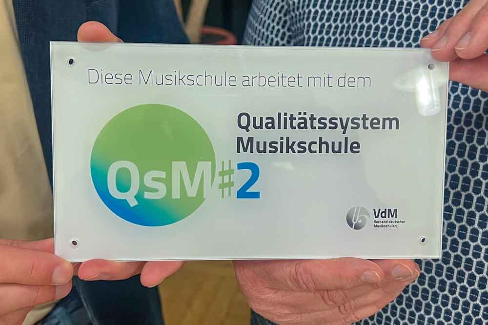 Stadtverwaltung Eine kleine Tafel wird in die Kamera gehalten, auf der steht u.a. Qualitätssystem Musikschule