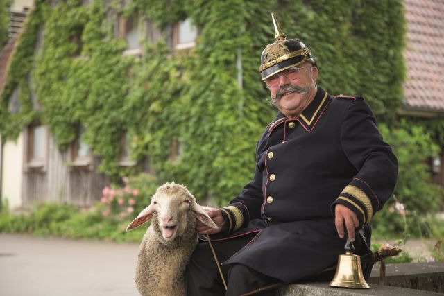 Der Büttel sitzt auf einem Stein, neben ihm der Hammel