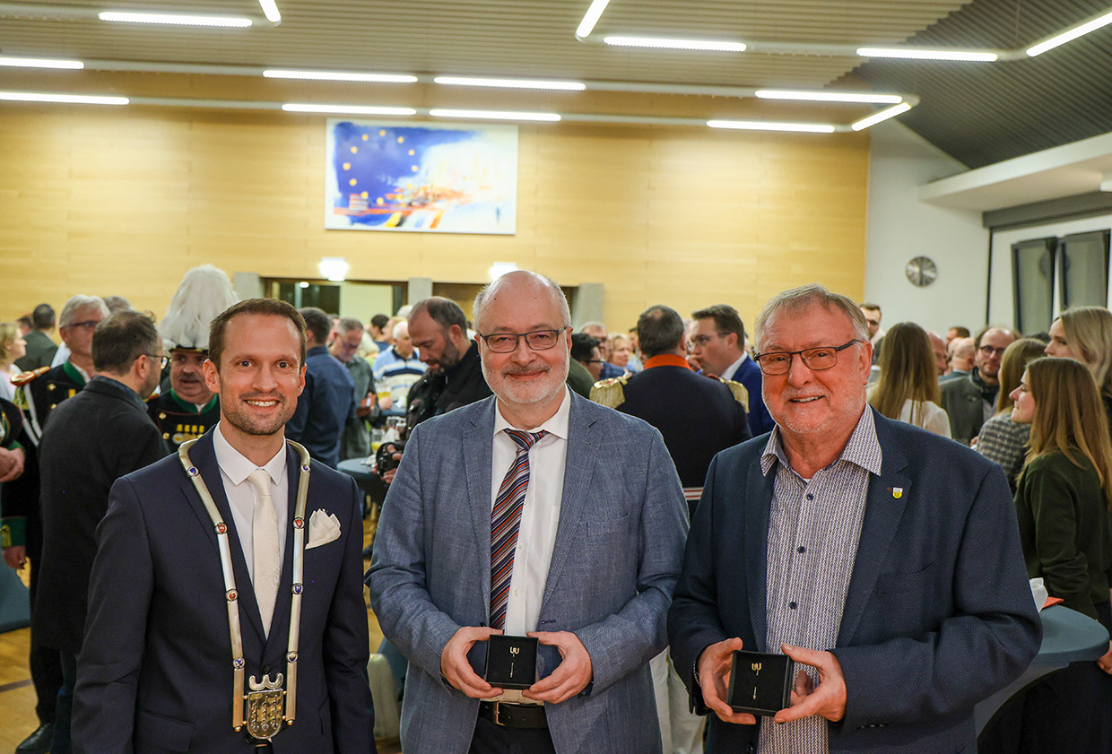 Drei Männer stehen in einem vollen Saal und lächeln in die Kamera