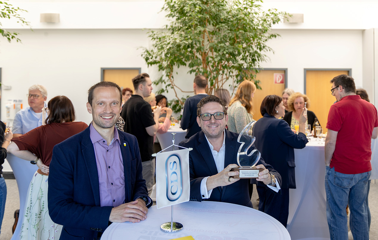 Zwei Männer stehen an einem Stehtisch udn lächeln in die Kamera, der rechte hebt eine Trophäe hoch