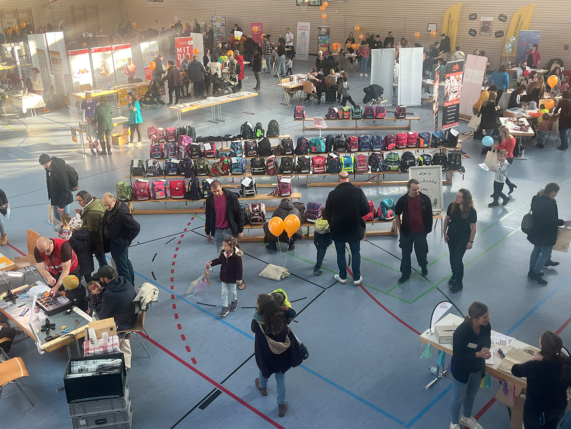 Viele Menschen in einer Turnhalle, in der viele Stände aufgebaut sind.