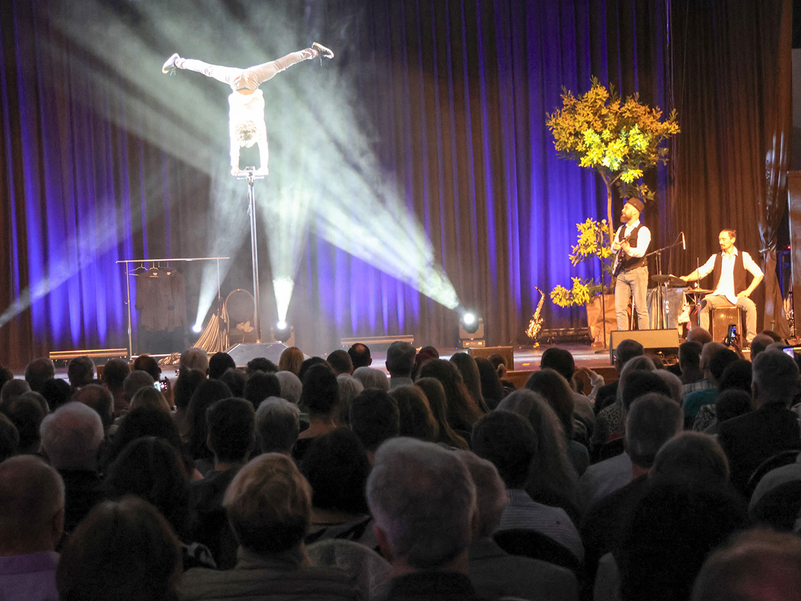 ein Akrobat macht Handstand auf einer Stange, daneben zwei Musiker