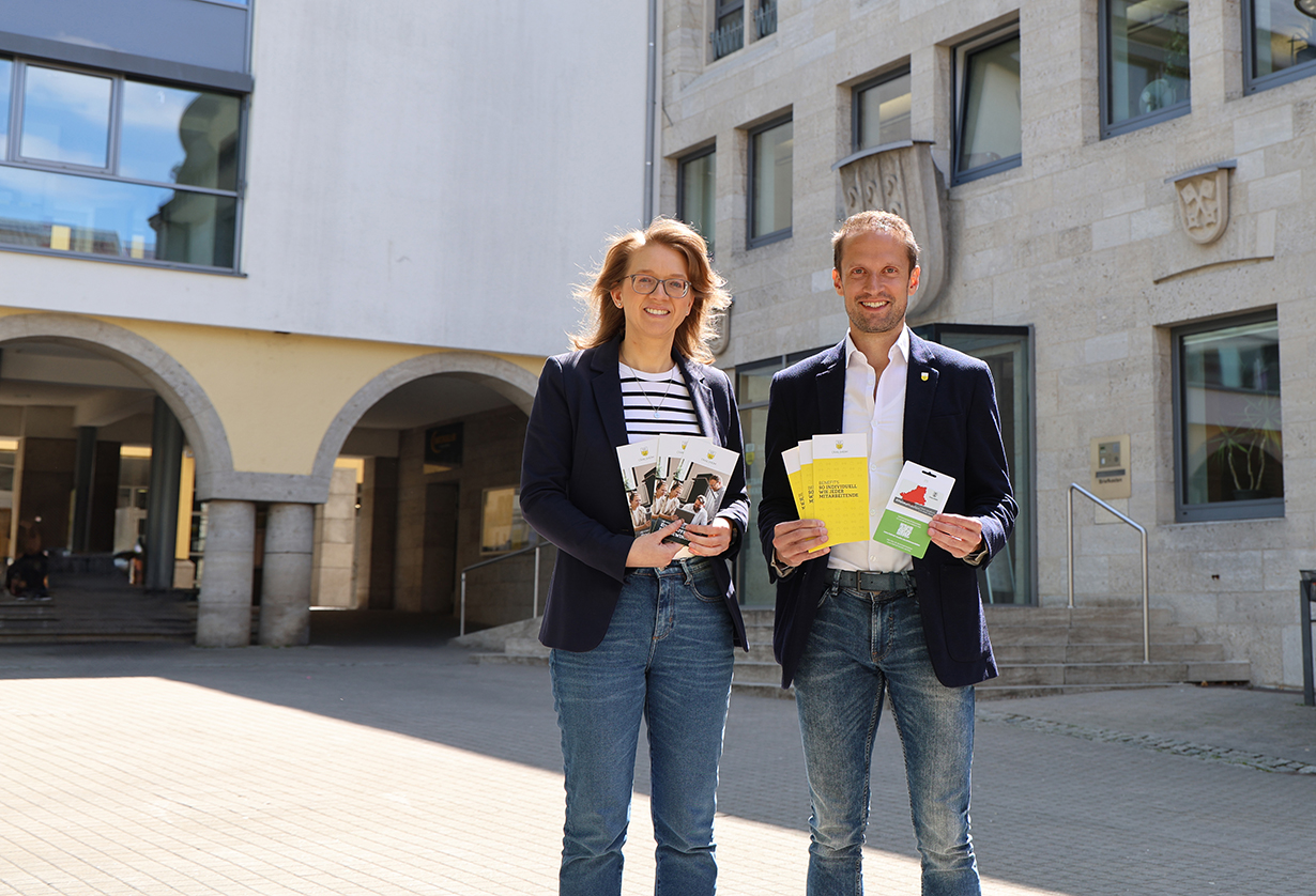 Eine Frau und ein Mann stehen vor einem großen Gebäude, halten Prospekte in der Hand und lächeln in die Kamera