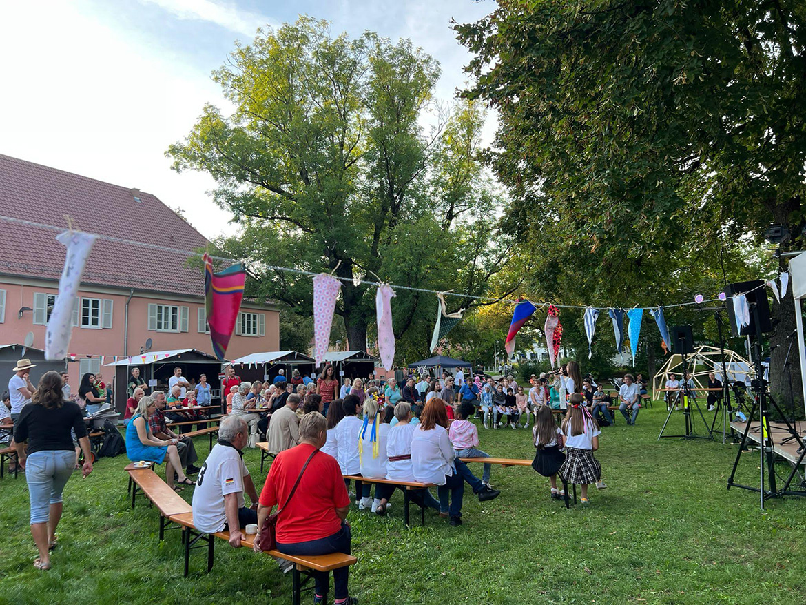 Viele Menschen sitzen in einem bunt geschmückten Garten auf Bänken und sehen einer Vorführung zu.