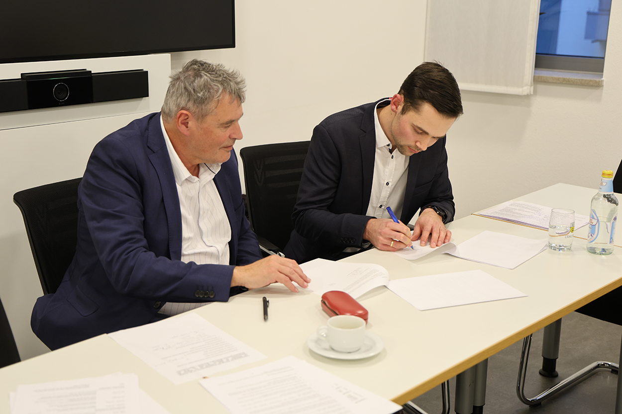 Ein älterer Herr sitzt an einem Tisch neben einem jüngeren Mann, der etwas unterschreibt