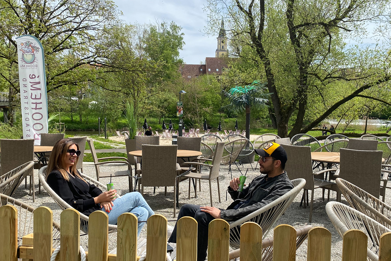 Eine Frau und ein Mann sitzen ganz realxt in einem gemütlichen Gästebereich, im Hintergrund sind ein Strand und eine Stadt zus ehen.