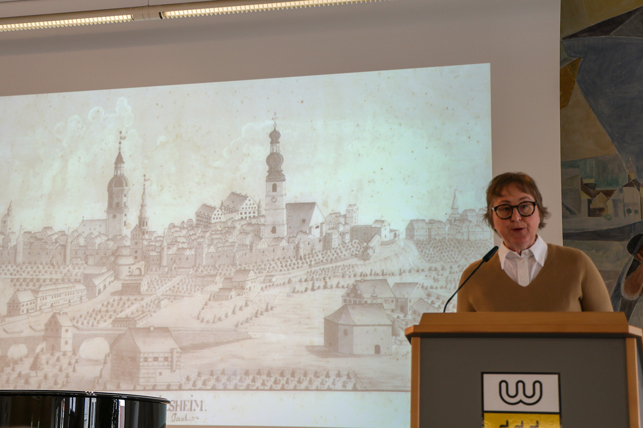 Stadtverwaltung Eine Frau steht an einem Rednerpult vor einer Leinwand auf der ein historisches Bild Crailsheims zu sehen ist.