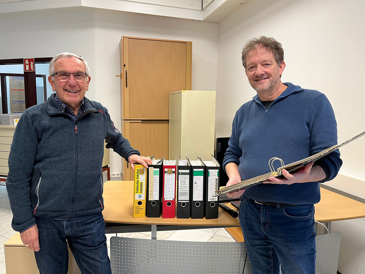 zwei Männer lächeln in die Kamera zwischen ihnen stehen auf einem Schreibtisch Ordner