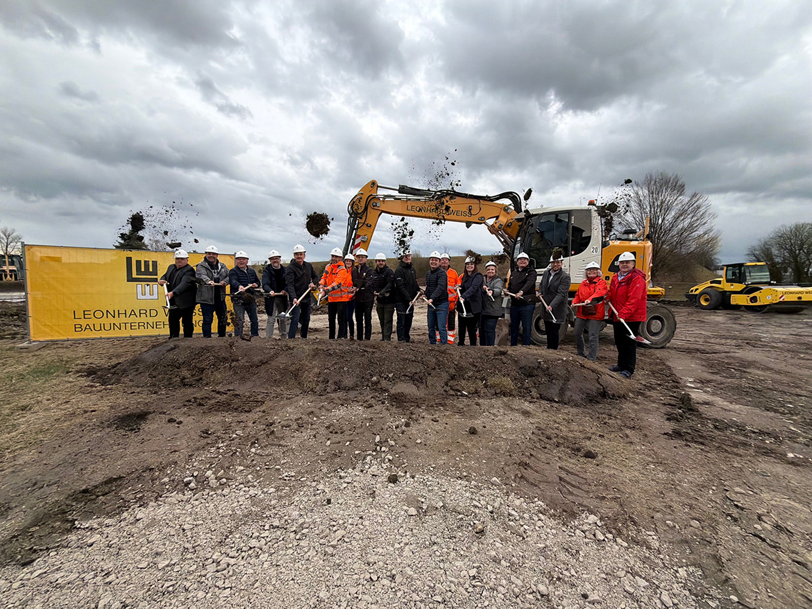 Viele Menschen mit Spaten und weißen Bauhelmen werfen vor einem Bagger Erde in die Luft